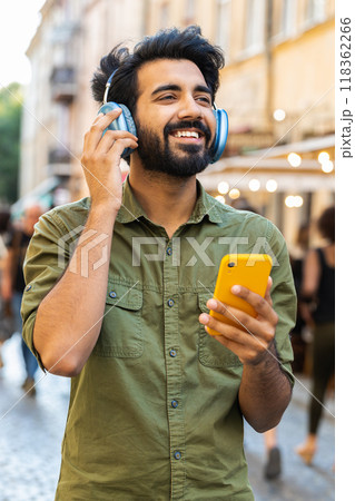 Happy young man in wireless headphones choosing, listening music dancing outdoors city street 118362266