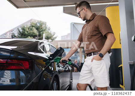 Low angle of concentrated young bearded male driver in sunglasses and casual clothes filling fuel into modern car with gas pump nozzle at station Low angle of concentrated young bearded male driver in sunglasses and casual clothes filling fuel into modern car with gas pump nozzle at station 118362437