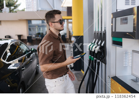 Handsome young man refueling his luxury car and looking on the scoreboard while standing on self service gas station Handsome young man refueling his luxury car and looking on the scoreboard while standing on self service gas station 118362454