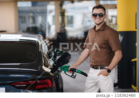 Smiling man refuelling his luxury car at the gas station. Guy looking camera and pouring petrol into tank of modern vehicle on filling station in city Smiling man refuelling his luxury car at the gas station. Guy looking camera and pouring petrol into tank of modern vehicle on filling station in city 118362460