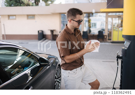 Handsome young man wipes his hands with a wet napkin after refuelling car on gas station. Casual style. Luxury car Handsome young man wipes his hands with a wet napkin after refuelling car on gas station. Casual style. Luxury car 118362491