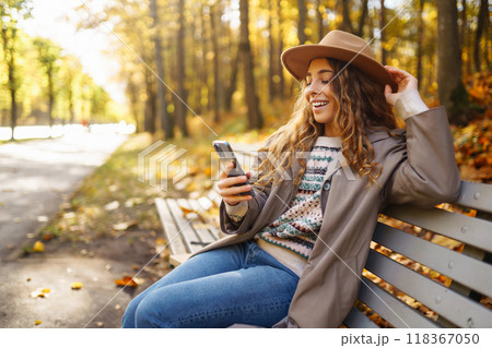 Young smiling woman sitting on bench in autumn park using phone in stylish clothes happy mood. 118367050
