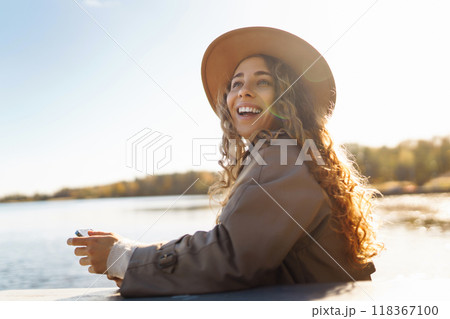 Smiling woman in hat on terrace with phone enjoys sunny autumn weather. Concept of relaxation, rest. Smiling woman in hat on terrace with phone enjoys sunny autumn weather. Concept of relaxation, rest. 118367100