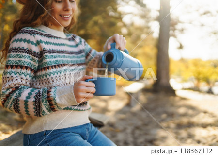 Woman holds thermos and drinks tea in sunny autumn forest. Vacation, travel, lifestyle concept. 118367112