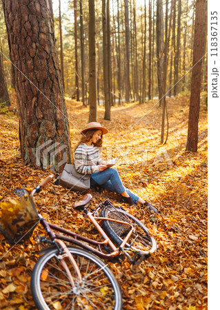 Stylish woman with a book near tree enjoys autumn weather in the park on the background of the bike. 118367135