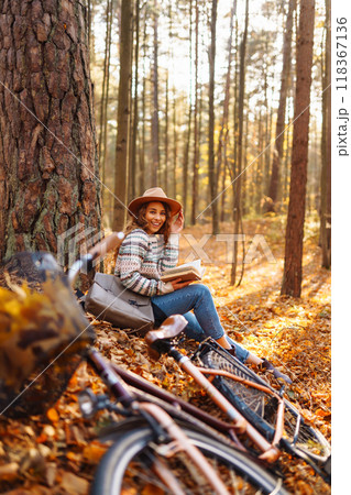 Stylish woman with a book near tree enjoys autumn weather in the park on the background of the bike. Stylish woman with a book near tree enjoys autumn weather in the park on the background of the bike. 118367136
