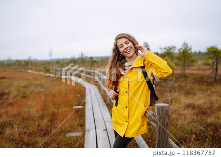 Smiling female traveler in yellow coat walks along wooden path among wild nature. Travel, vacation. 118367787