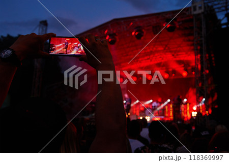 Person standing in front of stage, recording live music concert on smartphone. People crowd near stage enjoys musician band's performance Person standing in front of stage, recording live music concert on smartphone. People crowd near stage enjoys musician band's performance 118369997