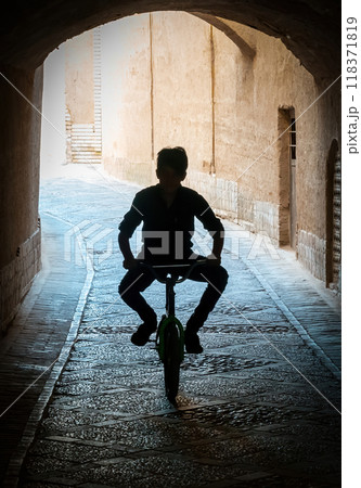 Silhouette of a kid riding a bicycle through a dimly lit tunnel in the narrow streets of the ancient city of Yazd Silhouette of a kid riding a bicycle through a dimly lit tunnel in the narrow streets of the ancient city of Yazd 118371819