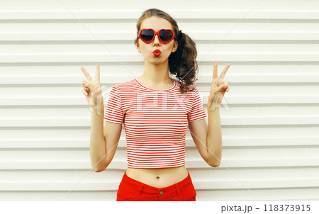 Beautiful young woman blowing kiss wearing red heart shaped sunglasses on white wall background 118373915