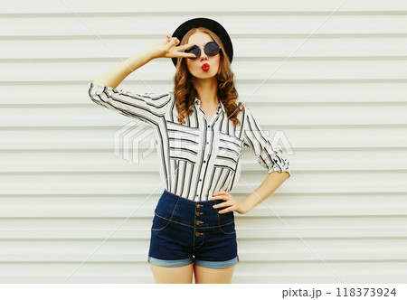 Attractive young woman blowing kiss in black round hat, shorts, striped shirt on white background Attractive young woman blowing kiss in black round hat, shorts, striped shirt on white background 118373924
