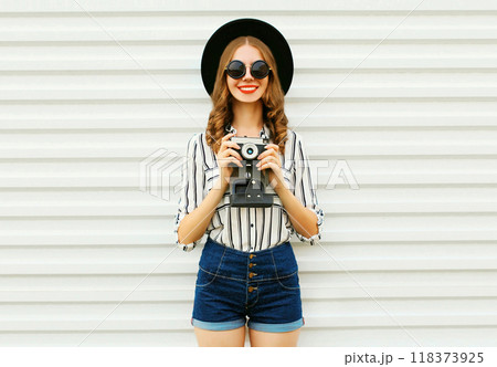 Happy smiling young woman holding camera in black hat, white striped shirt on white wall background Happy smiling young woman holding camera in black hat, white striped shirt on white wall background 118373925