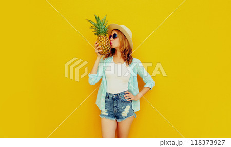 Beautiful young woman kissing pineapple in summer straw hat, shorts on colorful yellow background Beautiful young woman kissing pineapple in summer straw hat, shorts on colorful yellow background 118373927