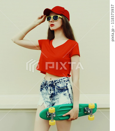 Summer portrait of young woman with green skateboard wearing a shorts and red baseball cap in a city Summer portrait of young woman with green skateboard wearing a shorts and red baseball cap in a city 118373937