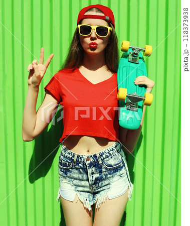 Summer portrait of young woman with green skateboard wearing a shorts and red baseball cap in a city Summer portrait of young woman with green skateboard wearing a shorts and red baseball cap in a city 118373938