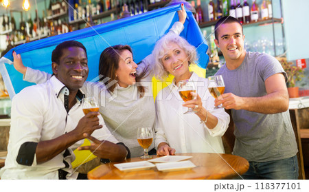 Excited male and female football fans celebrating championship win, waving flag of Ukraine in pub Excited male and female football fans celebrating championship win, waving flag of Ukraine in pub 118377101