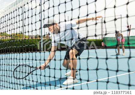 Portrait of man playing with male partner tennis doubles game on outdoors court. View through tennis net 118377161