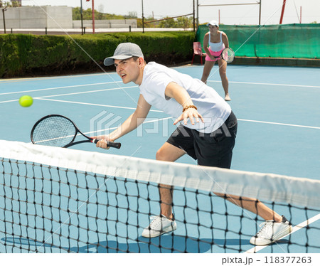 Portrait of young guy playing tennis on open court in summer, swinging racket to return ball over net. Sportsman ready to hit volley Portrait of young guy playing tennis on open court in summer, swinging racket to return ball over net. Sportsman ready to hit volley 118377263