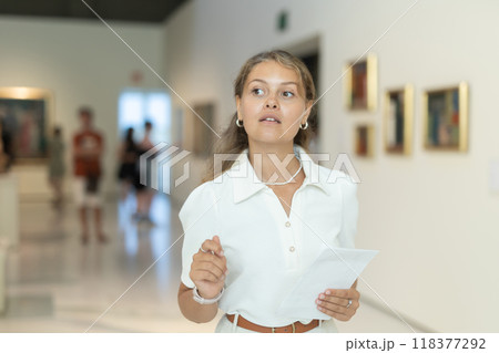 Young girl holding an information booklet looks at paintings in an art exhibition in museum 118377292