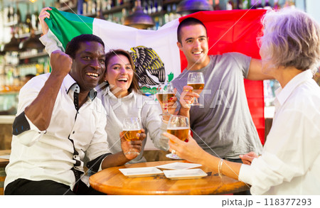 Excited multiracial soccer fans with flag of Mexico celebrating victory with pint of beer in the pub 118377293