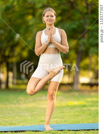 Girl in park on green grass of lawn performs Ardha Baddha Padma Vrikshasana Girl in park on green grass of lawn performs Ardha Baddha Padma Vrikshasana 118377307