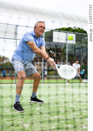 European old man holding playing padel during training in court. View through tennis net 118377332
