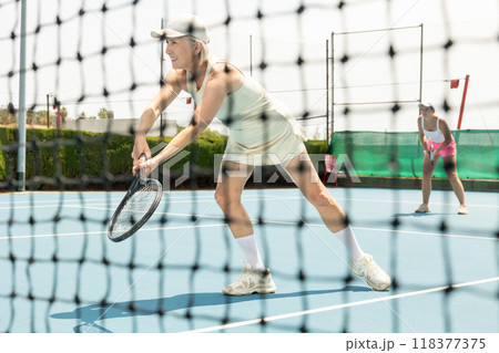 Active aged woman plays tennis on court. View through tennis net Active aged woman plays tennis on court. View through tennis net 118377375