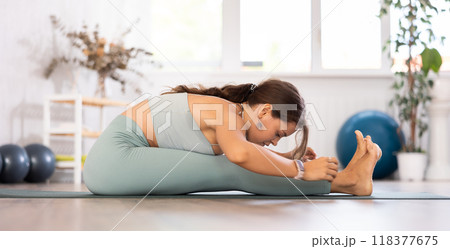 Young woman yoga trainer performing seated forward bend pose on black rug 118377675