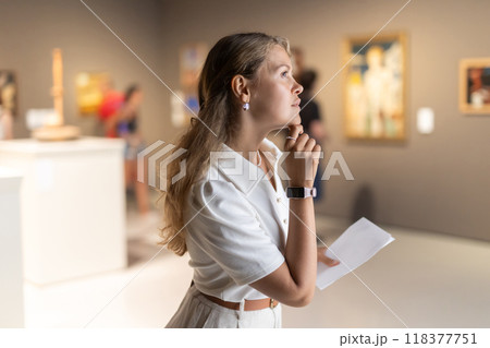 Girl visitor looks with interest at the sculpture in the museum hall, holding an information booklet in her hands 118377751