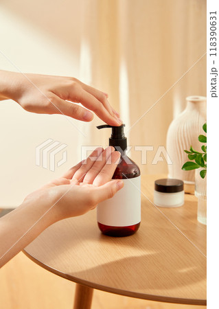A hand pumps a bottle of lotion, with a blank white label, onto another hand, placed on a wooden table with a vase of branches, a small jar of cream in the beige background suitable for skincare ads. A hand pumps a bottle of lotion, with a blank white label, onto another hand, placed on a wooden table with a vase of branches, a small jar of cream in the beige background suitable for skincare ads. 118390631