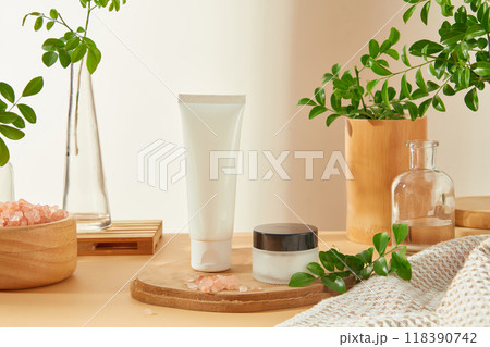 A minimalist beauty product promotion template features a white tube and jar are featured on a wooden platform with pink salt bowl, branches in a glass bottle, towel. The soft, natural light and warm. 118390742