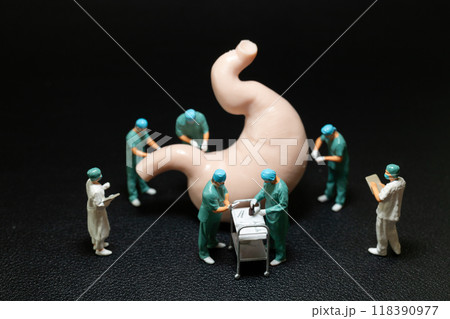 Miniature surgeons in scrubs performing an operation on a large model of a human stomach. 118390977
