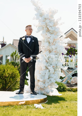 Elegant Groom in Black Tuxedo Standing by Stunning White Floral Arrangement at Outdoor Wedding Ceremony Elegant Groom in Black Tuxedo Standing by Stunning White Floral Arrangement at Outdoor Wedding Ceremony 118394703