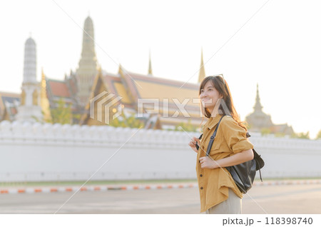 Traveler asian woman in her 30s, backpack slung over her shoulder, explores the intricate details of Wat Pra Kaew with childlike wonder. Sunlight dances on the golden rooftops. 118398740