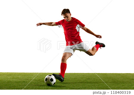 Young athletic man, football player, in red-white uniform, practicing powerful kick on green field against white studio background. Young athletic man, football player, in red-white uniform, practicing powerful kick on green field against white studio background. 118400319