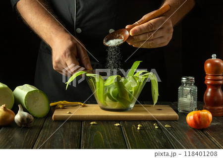 A cook adds salt with a spoon to vegetable marrow sliced into thin strips in a glass bowl. Concept of cooking tasty and healthy food with zucchini A cook adds salt with a spoon to vegetable marrow sliced into thin strips in a glass bowl. Concept of cooking tasty and healthy food with zucchini 118401208