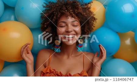 Happy birthday. A joyful woman with curly hair stands in front of blue balloons, dressed in an elegant summer dress and wearing colorful earrings. She has her hands raised as if celebrating something 118406747