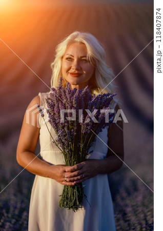 Blonde woman poses in lavender field at sunset. Happy woman in white dress holds lavender bouquet. Aromatherapy concept, lavender oil, photo session in lavender 118407874