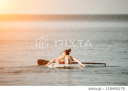 Sea woman sup. Silhouette of happy middle aged tanned woman in rainbow bikini, surfing on SUP board, confident paddling through water surface. Idyllic sunset. Active lifestyle at sea or river. 118408179