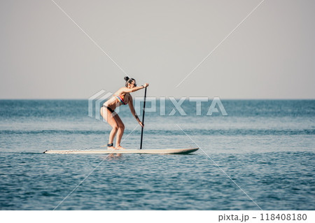 Sea woman sup. Silhouette of happy middle aged tanned woman in rainbow bikini, surfing on SUP board, confident paddling through water surface. Idyllic sunset. Active lifestyle at sea or river. 118408180