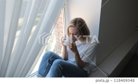 A woman drinks tea while sitting on the windowsill by the window. A woman in a white T-shirt admires the view from the window while sitting on the windowsill in her room and holding a cup. A woman drinks tea while sitting on the windowsill by the window. A woman in a white T-shirt admires the view from the window while sitting on the windowsill in her room and holding a cup. 118409348