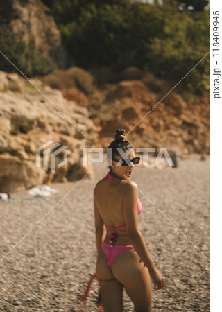 A woman in a bright pink bikini stands on a sunlit beach, enjoying her surroundings 118409946