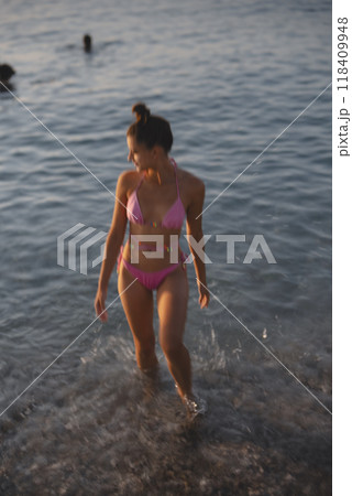 A woman gracefully walking in shallow water while wearing a pink bikini at the beach 118409948
