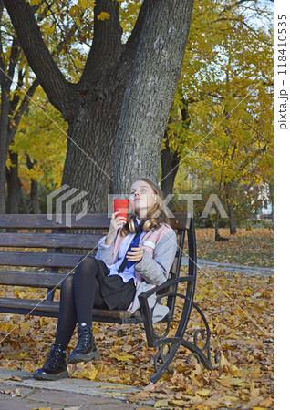 a girl sits on a bench in an autumn park, headphones on her head a girl sits on a bench in an autumn park, headphones on her head 118410535