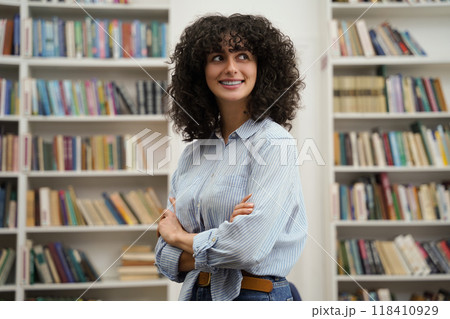 Curly-haired pretty young woman standing on the bookshelves background Curly-haired pretty young woman standing on the bookshelves background 118410929