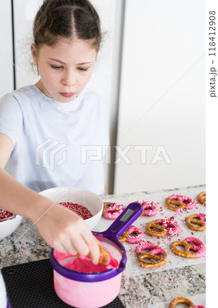 With focused attention, this budding culinary artist dips pretzels into a pot of melted chocolate, creating sweet delights in the warmth of a well-lit home kitchen. 118412908