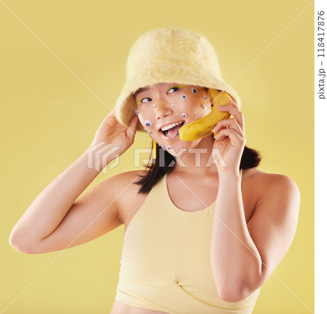 Woman, banana phone call and studio with smile, communication and eyes sticker art on face. Asian model girl, yellow background and fashion with beauty, aesthetic and cosmetic wellness with fruit 118417876