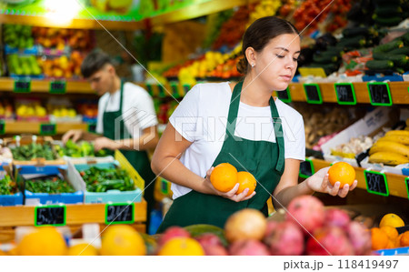 Hardworking young saleswoman puts fresh oranges on the counter Hardworking young saleswoman puts fresh oranges on the counter 118419497