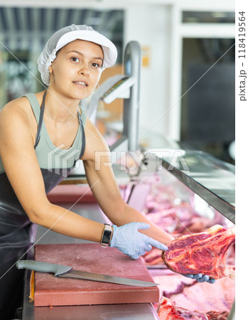 Girl in butcher shop cuts meat carcass into small pieces of meat for retail sale in small store Girl in butcher shop cuts meat carcass into small pieces of meat for retail sale in small store 118419564