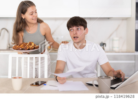 Busy guy working on notebook in the kitchen and a girl offering him pie Busy guy working on notebook in the kitchen and a girl offering him pie 118419628